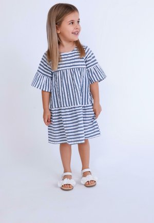 Robe rayée en bleu clair et blanc, avec des manches courtes évasées, une taille froncée et un ourlet superposé. Assortie à des sandales blanches.