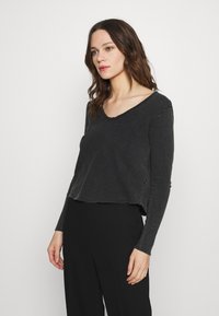 Haut noir à manches longues, court, avec un large décolleté et un ourlet brut. Le tissu semble doux avec une texture légèrement usée, assorti à un pantalon noir.