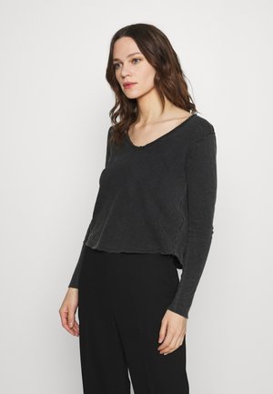 Haut noir à manches longues, court, avec un large décolleté et un ourlet brut. Le tissu semble doux avec une texture légèrement usée, assorti à un pantalon noir.