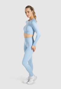 Hellblaues Sportset besteht aus einem langärmligen Crop-Top und hoch taillierten Leggings aus dehnbarem Stoff, kombiniert mit weißen Turnschuhen.