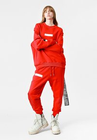 Sudadera roja y joggers a juego hechos de una tela suave. La sudadera cuenta con una etiqueta metálica; los joggers tienen bolsillos con cremallera y detalles en los laterales.