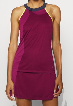 Femme portant une robe de sport sans manches de couleur magenta foncé avec col noir et bordure jaune, debout devant un fond uni.