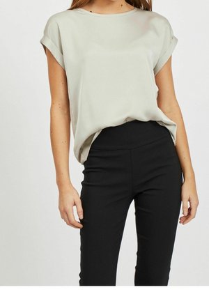 Frau trägt eine hellbeige kurzärmelige Bluse, die in schwarze, hoch taillierte, eng anliegende Hosen gesteckt ist, dargestellt vom Hals bis zu den Hüften.