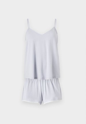 Jeune femme debout, pieds nus, portant un haut sans manches bleu clair et un short assorti sur fond blanc uni.