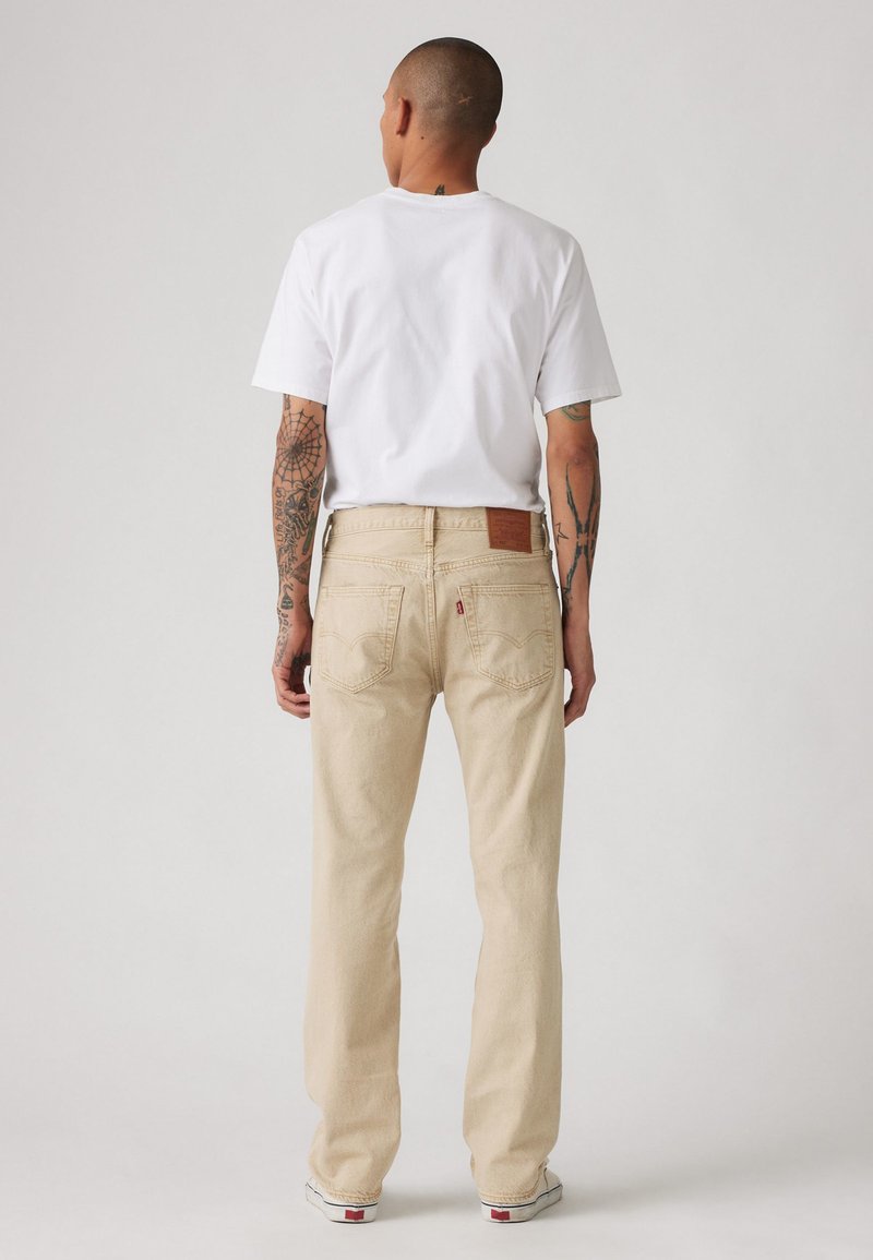 Beige gerade geschnittene Jeans mit einem braunen Lederpatch; getragen mit einem schlichten weißen T-Shirt und weißen Sneakers. Sichtbare Tattoo-Details an den Armen.