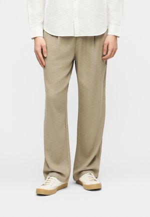 Személy bézs, bő szárú nadrágot, fehér gombos inget és fehér, gumitalpú tornacipőt visel, egyszerű háttér előtt áll.