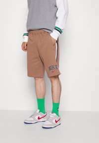 Person som bär en grå sweatshirt och bruna shorts med "Nike"-logotyp, kombinerat med klara gröna strumpor och grå Nike-sneakers.