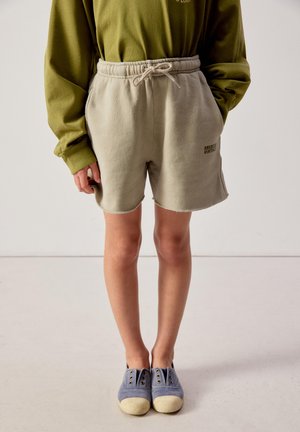 Bambino che indossa pantaloncini beige con coulisse con il logo "American Vintage", maglietta a maniche lunghe color oliva e scarpe slip-on blu, in piedi su un pavimento chiaro.