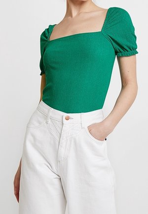 Mujer con camiseta verde de escote cuadrado con mangas abullonadas y pantalones blancos de cintura alta, con una mano en el bolsillo, sobre un fondo liso.
