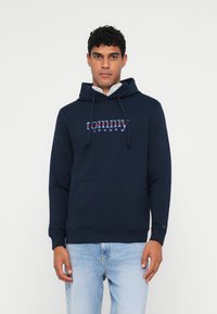 Navy hoodie featuring a kangaroo pocket, drawstring hood, and 'tommy jeans' logo in red and white. Made of soft, textured fabric.