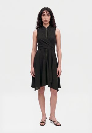 Une femme porte une robe portefeuille noire sans manches à jupe asymétrique et des escarpins noirs à brides, sur un fond blanc uni.