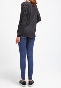 Woman wearing a loose black long-sleeve top, blue skinny jeans, and black slip-on shoes, standing with her back to the camera.