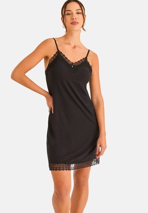 Vrouw draagt een zwarte mouwloze jurk met kanten rand aan de halslijn en zoom, poseert met één hand op de heup tegen een effen achtergrond.