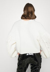 Weißer, gestrickter Pullover mit weiten Ärmeln, strukturierten Details und verkürztem Schnitt. Kombiniert mit glänzenden, schwarzen Hosen.