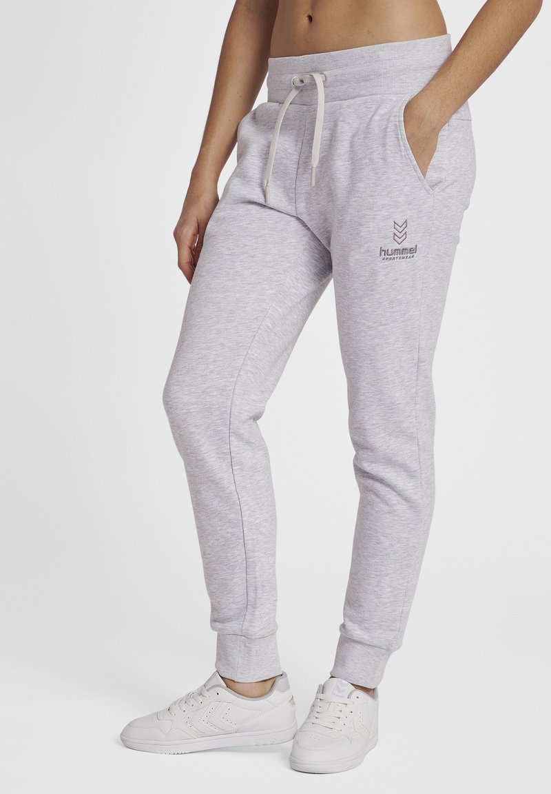Pantalon de survêtement slim gris clair avec taille à cordon, poches latérales, logo sur la cuisse, porté avec des baskets blanches sur une personne debout.