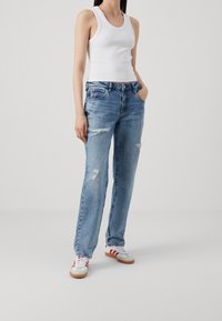 Jeans in denim blu chiaro con una vestibilità ampia, caratterizzati da dettagli sfrangiati e design a cinque tasche. Indossati con una canotta bianca e scarpe sportive.