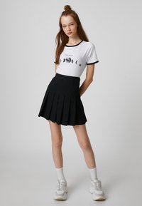 Biała koszulka z czarnymi akcentami i grafiką księżyca, zestawiona z czarną plisowaną spódnicą. Modelka w białych skarpetkach i masywnych sneakersach.