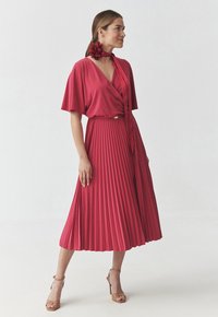 Abito plissettato di colore lampone con maniche a batwing corte, scollatura incrociata e vita cin cin. Completato con un accessorio floreale coordinato.