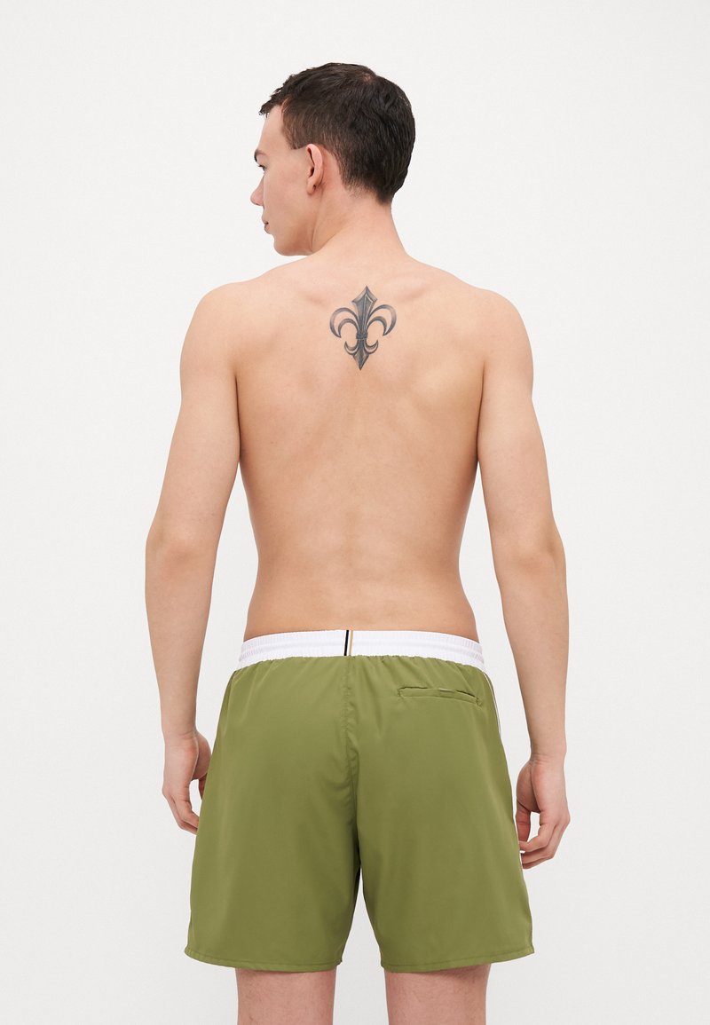 Pantaloni da bagno verdi con cintura elastica bianca, dotati di una tasca sul retro e una texture liscia. Il modello ha i capelli corti e un tatuaggio.