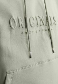 Sudadera verde claro con el texto "ORIGINALS" en relieve, cordones con puntas metálicas y un bolsillo tipo canguro. Tejido de algodón texturizado.