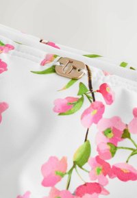 Material alb cu model floral roz și verde, având o etichetă mică dreptunghiulară aurie cu litera „b” decupată, atașată cu un fir alb.