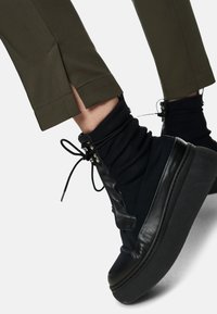 Black high-top shoes with a soft textile upper and leather accents, featuring a thick black sole and adjustable laces. Olive green pant hem is visible.