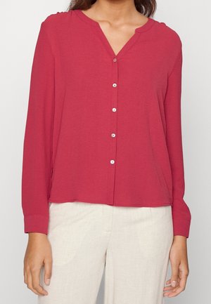 Femme portant une blouse rouge à manches longues boutonnée, associée à un pantalon beige clair sur un fond uni.