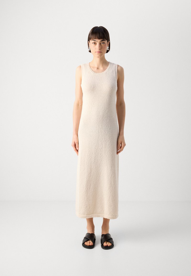 Robe crème sans manches, longueur genou avec un tissu texturé. Le design présente un col rond et une coupe droite, assortie à des sandales noires.