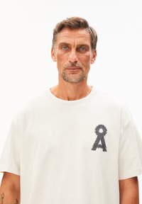 Witte katoenen t-shirt met een getextureerd zwart geborduurd ontwerp van een bewustwordingslint op de linker borst. Korte mouwen en ronde hals.