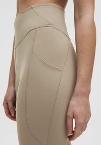 Magas derekú, bézs leggings sima, rugalmas anyagból, varrási részletekkel és egy oldalsó zsebbel, amelyen apró perforációk találhatók.