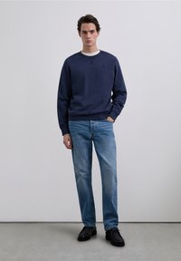 Marineblå sweatshirt med rund hals og ribbede manchetter, kombineret med lyseblå jeans. Modellen har sorte sko på og står foran en neutral baggrund.