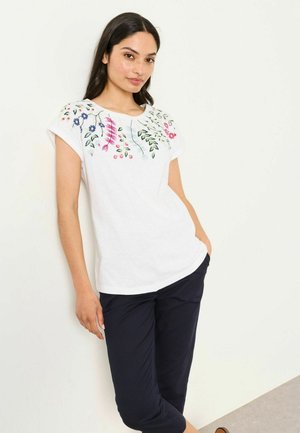 Junge Frau lehnt an einer weißen Wand, trägt ein weißes T-Shirt mit floralem Stickmuster und dunkelblaue knöchellange Hose, eine Hand in der Tasche.