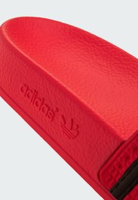 Rød skoslide med struktureret syntetisk materiale, præget branding og et strømlinet, buet design. Sort accentdetalje synlig på siden.
