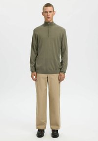 Haut vert olive clair à manches longues avec un col à demi-fermeture éclair ; texture lisse, ajusté à la taille, associé à un pantalon beige droit.