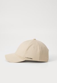 Beige baseballlippu, joka on valmistettu puuvillatwillistä. Siinä on kaareva lippa, kuusi paneelia ja "Calvin Klein"-logo brodeerattuna sivulle.
