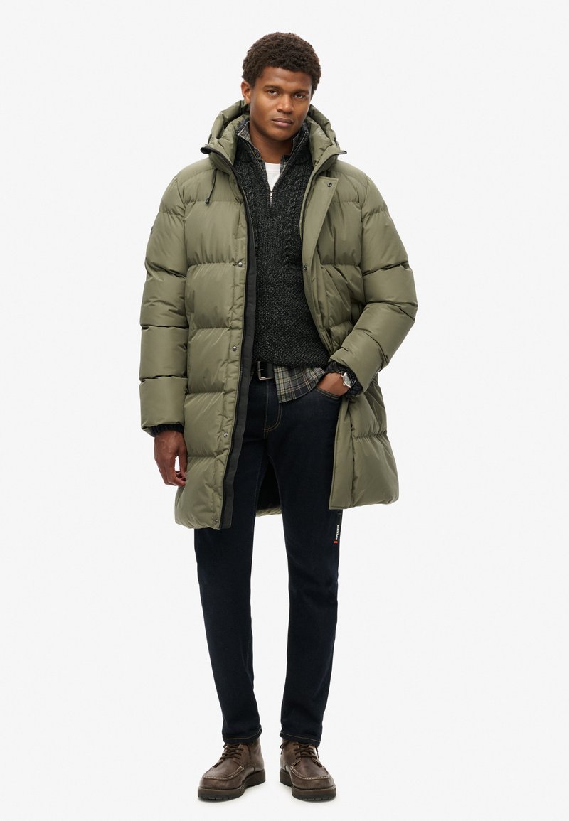 Abrigo acolchado verde con un cuello alto, sobre un suéter oscuro y una camisa a cuadros. Jeans oscuros y botas de cordones marrones complementan el atuendo.