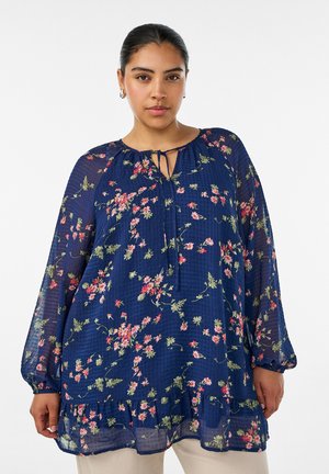 Kvinde med mørkt hår sat tilbage bærer en mørkeblå blomstret bluse med lange ærmer, bindebånd ved halsen og flæset kant.