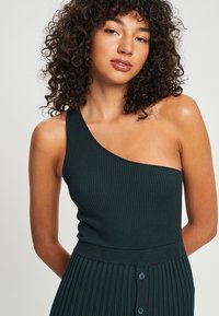 Ein schulterfreies, geripptes Kleid in dunkelgrün. Verfügt über ein tailliertes Oberteil und einen plissierten Rock mit Knopfdetails. Glatte Textur, minimales Design.