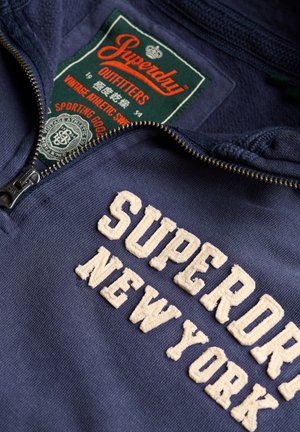 Marineblauwe sweatshirt met een ritskraag, voorzien van crèmekleurige geborduurde tekst "SUPERDRY NEW YORK" en een groene label erboven.