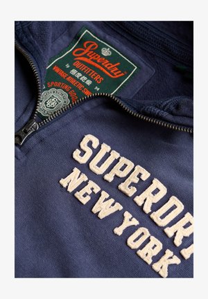 Marineblauer Pullover mit Reißverschluss-Kragen, verziert mit cremefarbenem, gesticktem Text "SUPERDRY NEW YORK" und einem grünen Etikett darüber.