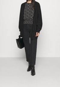Schwarzer oversized Cardigan über einem gemusterten schwarz-weißen Oberteil, kombiniert mit hochgeschnittenen schwarzen Jeans und Stiefeletten, während sie eine schwarze Handtasche hält.