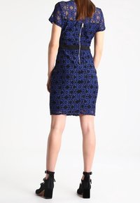 Žena v tmavomodrých krajkových šatech se květinovými vzory, které sahají po kolena a mají krátké rukávy, a v černých kotníkových botách s masivními podpatky.