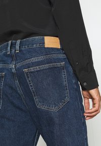 Jeans de mezclilla en un lavado azul oscuro con una etiqueta de cuero de WEEKDAY en la cinturilla. Presenta costuras en contraste y dos bolsillos traseros.