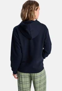 Haut à capuchon bleu marine avec une coupe décontractée, doté d'une capuche à cordon de serrage et de poignets et ourlet côtelés. Associé à un pantalon à carreaux verts.