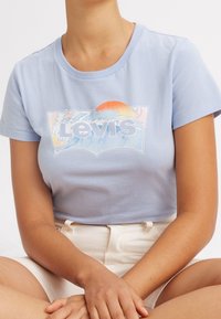 Osoba ubrana w jasnoniebieską koszulkę Levi's z grafiką fali i zachodu słońca, w połączeniu z białymi, wysokimi szortami, mająca ręce na kolanach.