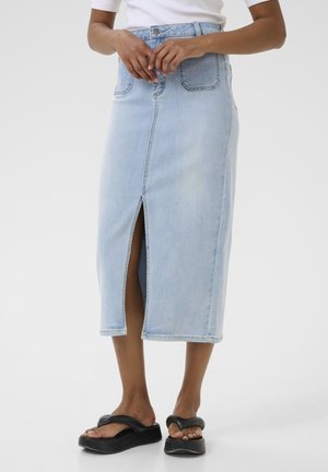 Jupe en denim bleu clair de coupe mi-longue, avec une fente à l'avant et deux poches arrière. Portée avec des sandales à plateforme noires.