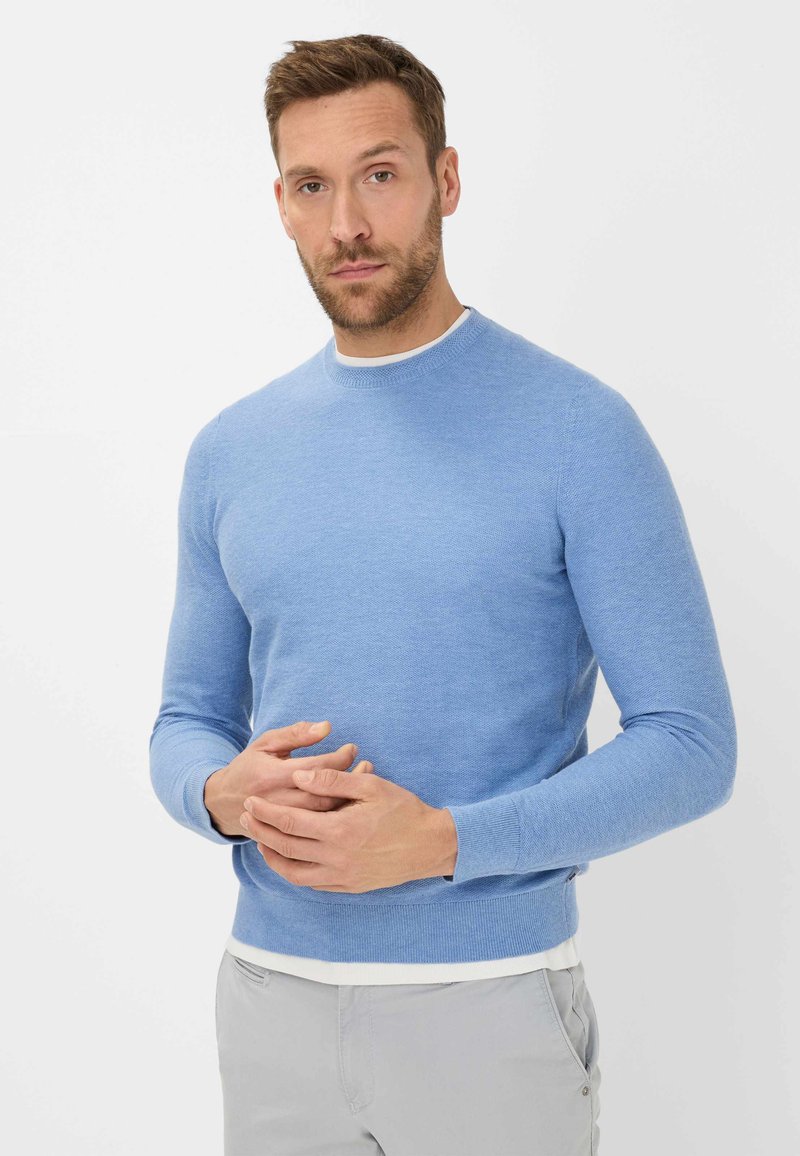 Hellblauer gestrickter Pullover mit runder Halslinie, langen Ärmeln und weicher Textur. Getragen über einem weißen Hemd, kombiniert mit hellgrauen Hosen.