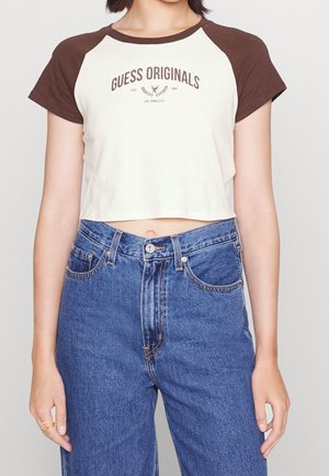 Nainen, joka käyttää valkoista lyhytmallista t-paitaa, jossa on ruskeat hihat ja teksti "GUESS ORIGINALS", yhdistettynä korkeavyötäröisiin sinisiin farkkuihin.