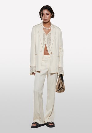 Un costume en lin couleur crème avec une veste cintrée, un pantalon à jambes larges et des détails en dentelle sur le haut. Accessoirisé avec un sac tissé texturé.