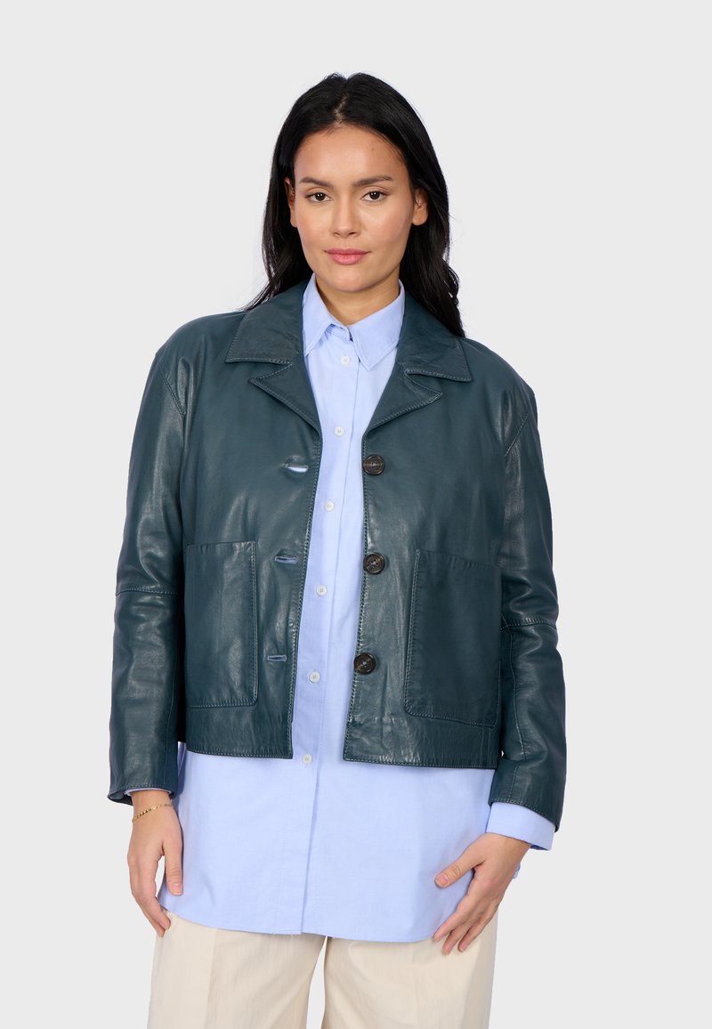 Zelená kožená zkrácená bunda s špičatým límcem, předními knoflíky, dvěma velkými kapsami a hladkým povrchem, nošená přes světle modrou košili.